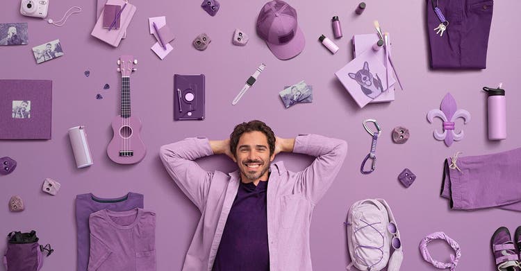 Smiling adult male with relaxed pose laying down on purple backdrop surrounded with various lifestyle items.